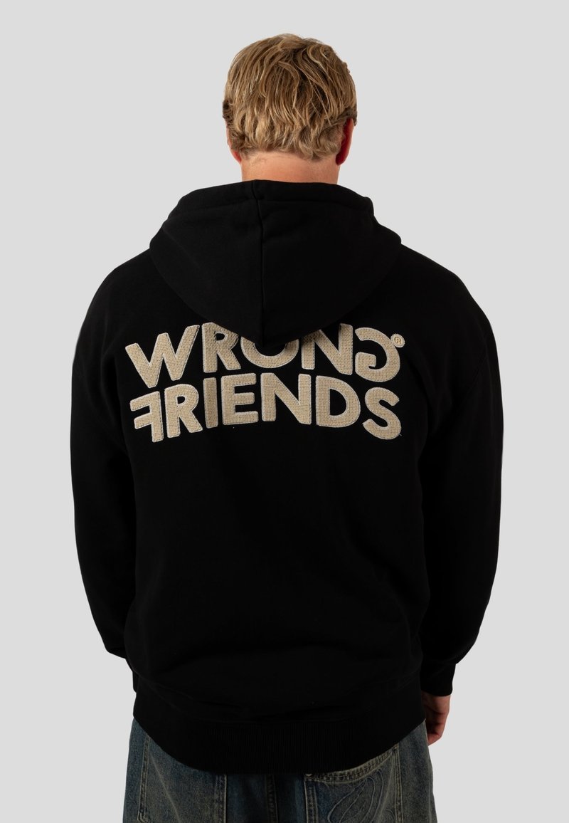 Černá mikina s kapucí, na zadní straně s velkým, texturovaným nápisem "WRONG FRIENDS" v světlé béžové barvě. Vyrobena z měkkého materiálu s uvolněným střihem.