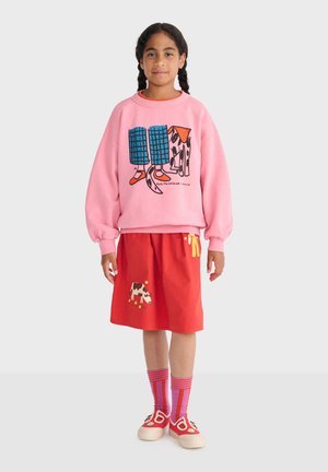 Fille aux cheveux tressés portant un pull rose avec un chien et un motif de jambes, une jupe rouge à imprimé vache, des chaussettes rayées et des chaussures rouges.