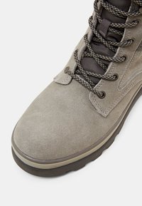 Botte de randonnée en daim gris clair avec des lacets noirs et gris à motif, des œillets cousus, une languette rembourrée et une semelle en caoutchouc épaisse.