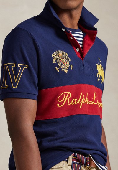 Mornarska polo majica s crvenim horizontalnim prugom, žutim natpisom "Ralph Lauren" i izvezenim logotipom. Ima preklopljeni ovratnik i kratke rukave.