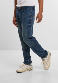 Donkerblauwe spijkerbroek met een relaxed fit, voorzien van voorsluiting, koperen klinknagels en een verwashed look. Gecombineerd met beige sneakers.