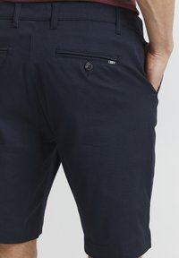 Shorts bleu marine en tissu texturé, avec une poche arrière dotée d'une fermeture à bouton et d'une petite étiquette décorative sur la poche.