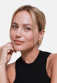 Pendientes de aro de oro llevados por una persona con cabello rubio claro, vestida con una blusa sin mangas de canalé negra, y con un maquillaje neutro.