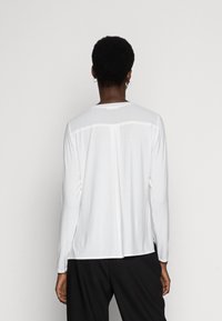 Haut blanc à manches longues avec un col rond, présentant une couture sur le dos et une coupe décontractée, fabriqué en tissu lisse et léger.