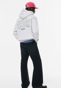 Felpa grigia con logo e l'anno '1991' sul retro, abbinata a jeans scuri. Gli accessori includono un cappellino rosso con stelle.