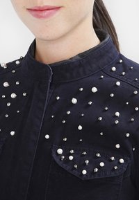 Marinblå denimskjorta med mandarin-krage, dekorerad med silver- och pärlliknande nitar över tyget och på bröstfickorna.