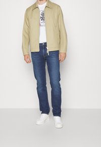 Světlá béžová bunda s špičatým límcem, zapínáním na zip a bočními kapsami, nošená přes bílé tričko s potiskem a modré džíny, doplněná bílými teniskami.