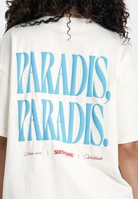 T-shirt surdimensionné blanc cassé avec un grand texte bleu "PARADIS, PARADIS." au dos, accompagné d'un petit détail rouge "Depuis 2008" et logo.