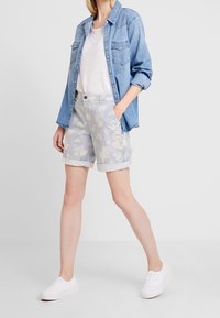 Een denim blouse over een wit T-shirt, gecombineerd met lichtblauwe shorts met bloemenpatroon en witte sneakers. De shorts hebben omgeslagen mouwen.