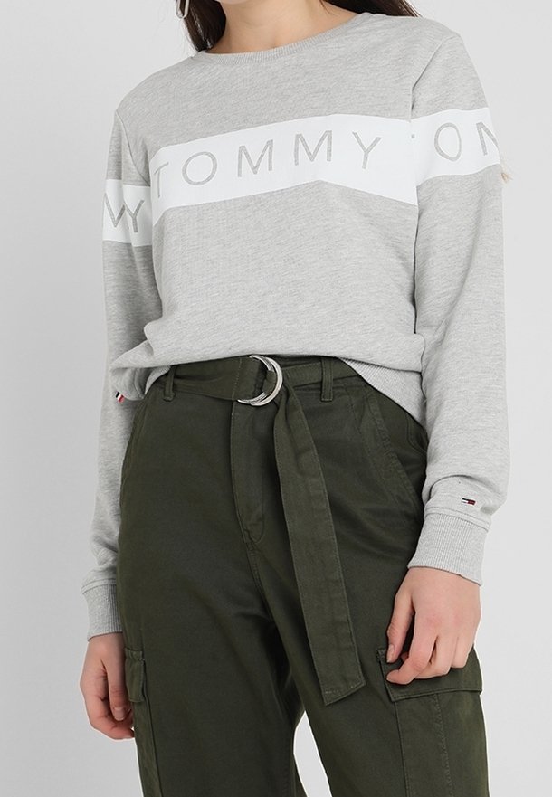 Šedá mikina s bílým vodorovným pruhem a nápisem "TOMMY". Nošena s olivově zelenými kalhotami, utaženým pásem a elastickými manžetami na zápěstích.