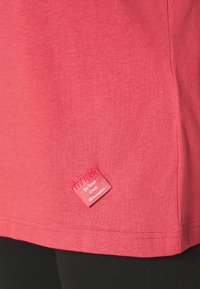 Gros plan sur un tissu rose corail avec une petite étiquette cousue portant l'inscription « Soyez votre propre champion » près de l'ourlet au-dessus d'un pantalon noir.