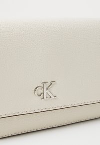 Cartera de piel blanca con un acabado texturizado, que presenta un logo "cK" en plata, acentos cosidos y un diseño minimalista.