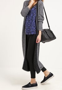 Lång grå stickad cardigan över en blåmönstrad topp och svarta leggings. Svarta slip-on skor. Svart handväska med axelrem.