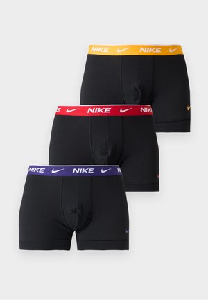 Mężczyzna stoi boso, ubrany w czarne bokserki Nike z czerwonym pasem, na jednolitym białym tle, ręce za plecami.