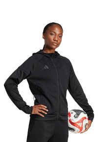 Felpa nera con zip, atletica e con finitura testurizzata, dotata di cappuccio e del logo Adidas sul petto. Il modello tiene in mano un pallone da calcio.