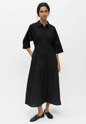 Frau trägt ein langes schwarzes Kleid mit Knopfleiste und Dreiviertelärmeln sowie schwarzen Schlupfschuhen, steht mit einer Hand in der Tasche.