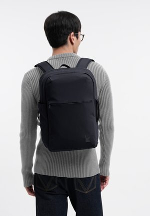 Henkilö, jolla on silmälasit, harmaa ribbineulepusero, tummat farkut ja musta suorakaiteen muotoinen reppu sivutaskuilla valkoisella taustalla.