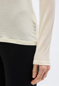 Κοντινό πλάνο ενός ατόμου που φοράει εκρού μακρυμάνικο πουκάμισο με το λογότυπο «icebreaker» κοντά στο τελείωμα και μαύρο παντελόνι, με το χέρι χαλαρό στο πλάι.