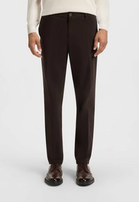 Pantalons marron foncé à coupe ajustée, avec une texture lisse, des plis sur le devant et une fermeture à bouton unique. Portés avec des chaussures en cuir marron.