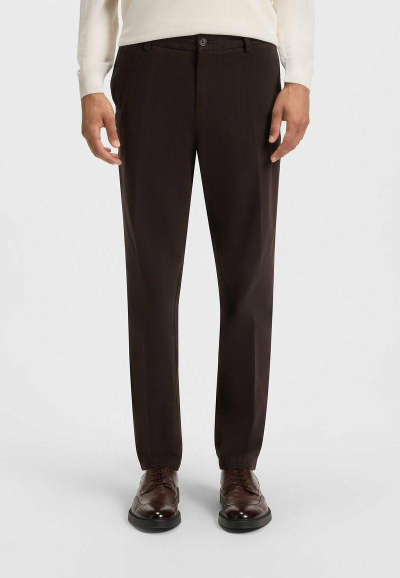 Pantalons marron foncé à coupe ajustée, avec une texture lisse, des plis sur le devant et une fermeture à bouton unique. Portés avec des chaussures en cuir marron.