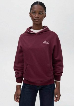 Frau in einem kastanienbraunen Kapuzenpullover mit dem Text "Fast Wochenende", dunklen Jeans und einem weißen Langarmshirt darunter, vor einem schlichten Hintergrund stehend.