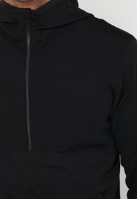 Haut à capuche noir zippé fabriqué en tissu texturé, avec une capuche et un zip partiel. Il dispose d'une poche avant et de coutures plates.