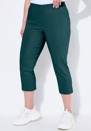 Kobieta ubrana w ciemnozielone spodnie o długości do połowy łydki i białe trampki, stojąca z jedną ręką opartą na biodrze na białym tle.