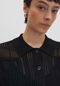 Schwarze Strickjacke mit einem Netzmuster, ausgestattet mit einem spitzen Kragen und zwei schwarzen Knöpfen, die ein strukturiertes Design zeigt.