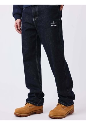 Personne portant un jean bleu foncé avec le logo "PXP" sur la cuisse et des bottes de travail marron clair sur fond blanc.