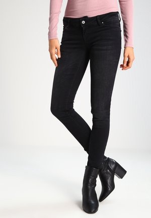Frau trägt schwarze Skinny-Jeans, schwarze Stiefeletten mit Blockabsatz und ein hellrosa geripptes Langarmshirt, steht mit überkreuzten Beinen.
