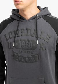 Felpa grigia con cappuccio e yoke nero, caratterizzata da una stampa del logo testurizzata e lacci. Tasca a marsupio frontale, design casual.
