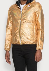 Zlatá metalická bunda na zip s kapucí, s elastickými manžetami a vícebarevným zipem. Nositelná přes bílé tričko a černé kalhoty.