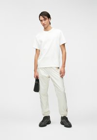 Λευκό βαμβακερό μπλουζάκι συνδυασμένο με κρεμ παντελόνι ελαφρού βάρους. Το μοντέλο κρατά μια μαύρη τσάντα, φορώντας μαύρα αθλητικά παπούτσια με σχέδιο. Ουδέτερο φόντο.