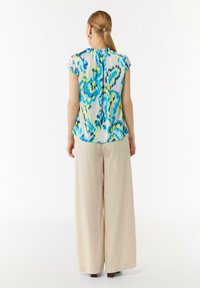 Mehrfarbige Bluse mit blauen, grünen und gelben Mustern. Kurzarm, plissierter Rücken, getragen mit beigen, weit geschnittenen Hosen. Glattes Material.