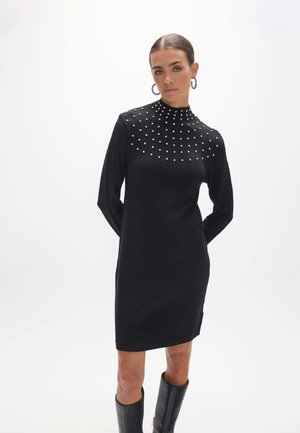 Mujer con vestido negro de manga larga con tachuelas plateadas en la parte superior del pecho y botas negras hasta la rodilla, de pie con las manos detrás de la espalda.