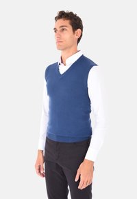 Gilet blu lavorato a maglia con scollatura a V, indossato sopra una camicia bianca con colletto, abbinato a pantaloni neri. Texture liscia e forma aderente.