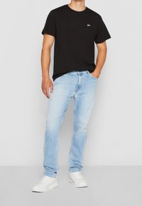 Musta lyhythihainen t-paita, jossa on pieni logo rinnassa, yhdistettynä vaaleansineisiin suoriin farkkuihin ja valkoisiin tenniskenkiin.