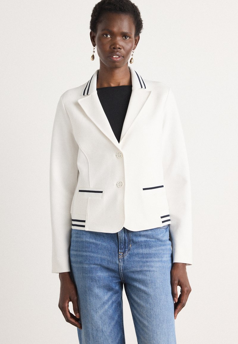 Blazer blanc à bouton unique, avec des accents rayés bleu marine sur le col et les poches, associé à un jean bleu. Tissu lisse, coupe ajustée.