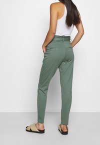 Pantalons verts ajustés avec taille haute, passants de ceinture et deux poches arrière. Portés avec un débardeur blanc et des sandales à glissière beiges.