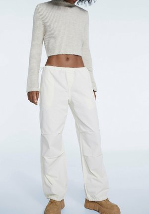 Femme portant un pull court en maille crème, un pantalon blanc ample et des chaussures à semelles épaisses couleur tan, debout devant un fond clair uni.