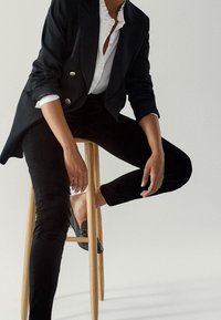 Svart dubbelknäppt kavaj, vit skjorta med krage, svarta figursydda byxor, sittande på en träpall, avslappnad handposition, eleganta svarta skor.