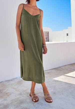 Elegantes olivengrünes Kleid mit V-Ausschnitt, dünnen Trägern und besticktem Saum, kombiniert mit braunen Riemchensandalen auf einer hellen Steinoberfläche.