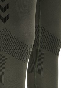Donkergroene leggings van rekbaar materiaal, met gestructureerde patronen en strepen in verschillende zwarttinten langs de zijkanten en knieën.