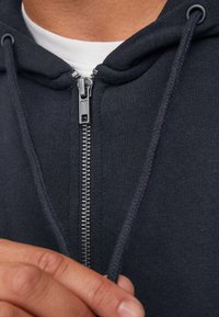 Haut à capuche zippé gris foncé avec une texture tricot à picots, fermeture éclair en métal et cordons de serrage. Comprend une capuche attachée pour plus de chaleur.