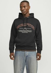 Sudadera negra con bolsillo frontal, que presenta texto en rojo y blanco en el pecho. Material de algodón, puños y dobladillo acanalados. Combinada con jeans de denim claros.