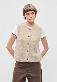 Bežna puhaste telovnik z okroglim izrezom, s 6 zlatimi gumbi spredaj, nošen preko belega T-shirta in v kombinaciji z rjavimi hlačami.