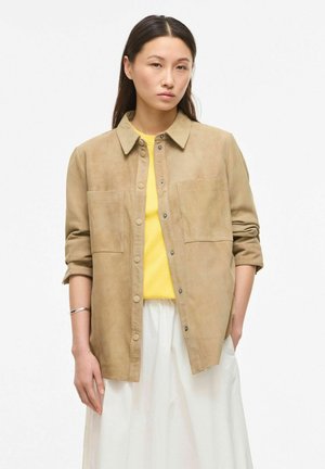 Jauna moteris dėvi smėlio spalvos užsegamą švarką virš geltonos palaidinės ir balto sijono, stovinti su rankomis kišenėse prieš balto fono foną.