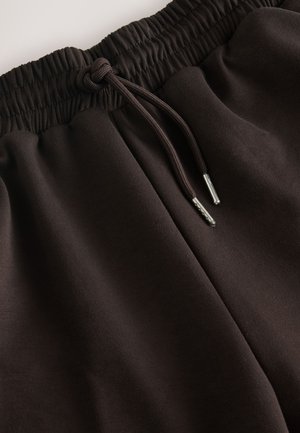 Pantaloni de trening maro, confecționați dintr-un material moale, cu talie elastică și șnur cu vârfuri metalice, prezentând o textură netedă și o croială lejeră.