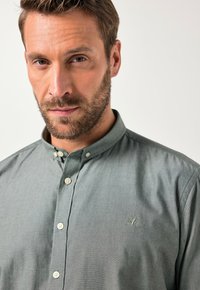 Camicia verdegrigia con bottoni e un piccolo emblema di un albero sul petto, caratterizzata da un colletto arrotondato e un tessuto testurizzato.