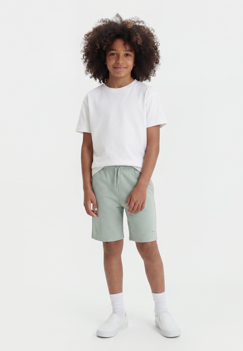 Enfant aux cheveux bouclés portant un t-shirt blanc, un short vert clair, des chaussettes blanches et des chaussures blanches à enfiler, debout devant un fond uni.